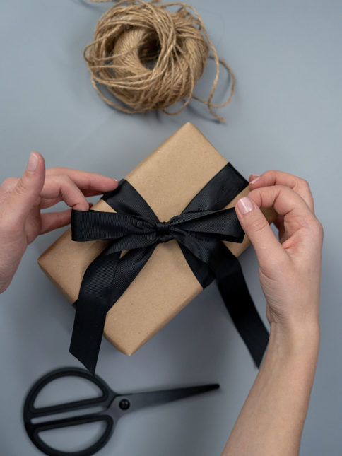 Zwei Hände ziehen die schwarze Schleife an einem nachhaltig verpackten Geschenk gerade. Darunter liegt eine Schere, drüber eine Rolle Garn.