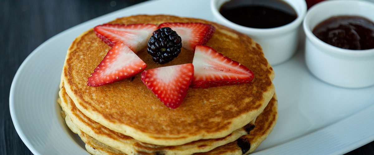 Ein Stapel Pancakes mit Erdbeeren und einer Brombeere, im Hintergrund Marmelade.