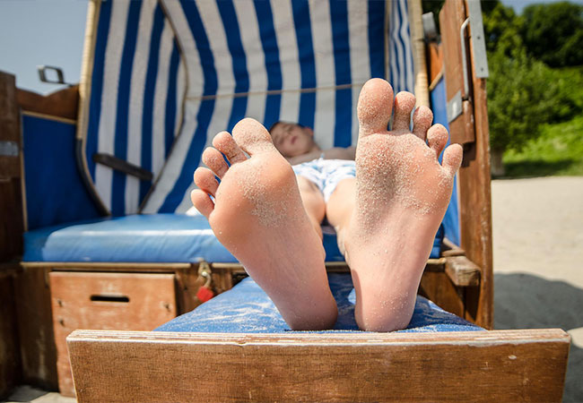 Im Mittelpunkt stehen die Füße eines Kindes. Das Kind schläft im Strandkorb Ostsee / Nordsee und hat Sand an den Füßen.