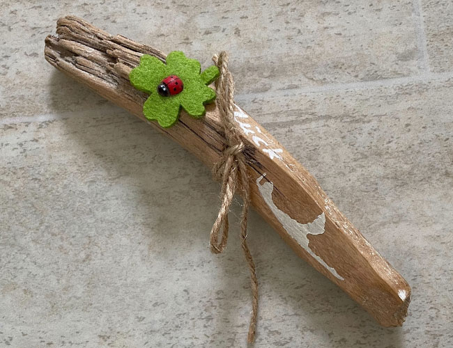 Ein kleines Stück Treibholz mit einer Schleife aus Garn und einem grünen Filz-Glückskleeblatt. Darauf sitzt ein hölzerner Marienkäfer. Das Holz wurde zusätzlich mit einer Sylt-Silhouette bemalt.