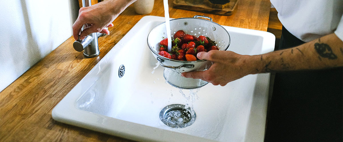 Die Hände eines Mannes halten eine Schüssel mit Erdbeeren, die er unter einem Wasserhahn abwäscht. Die Arbeitsplatte besteht aus Holz, das Spüle Material ist Keramik.