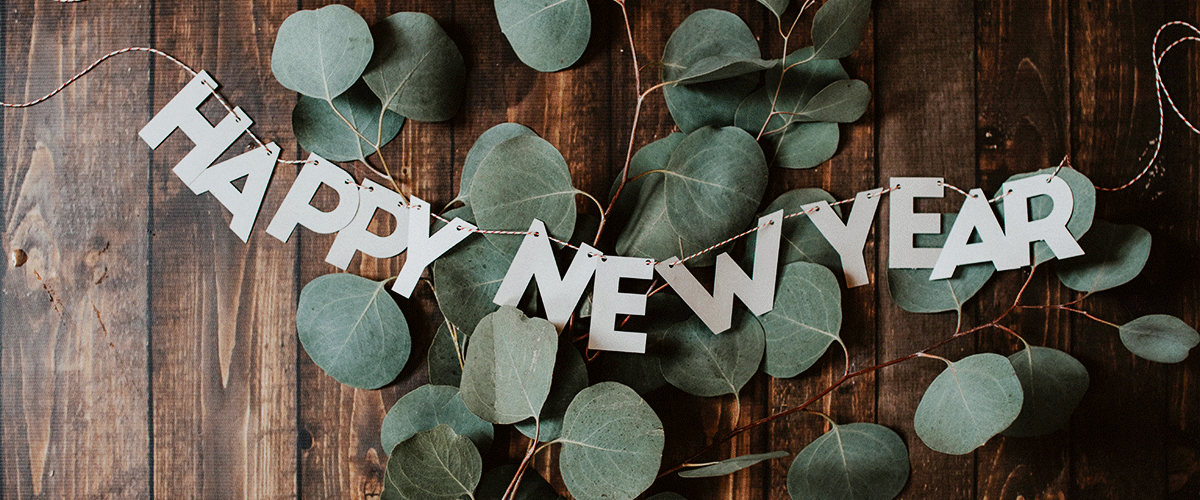 Auf einem braunen Holztisch liegen Eukalyptusblätter und auf eine Schnur aufgefädelte Buchstaben. Sie ergeben das Wort Happy New Year und sind somit die perfekte Silvester Tischdeko.