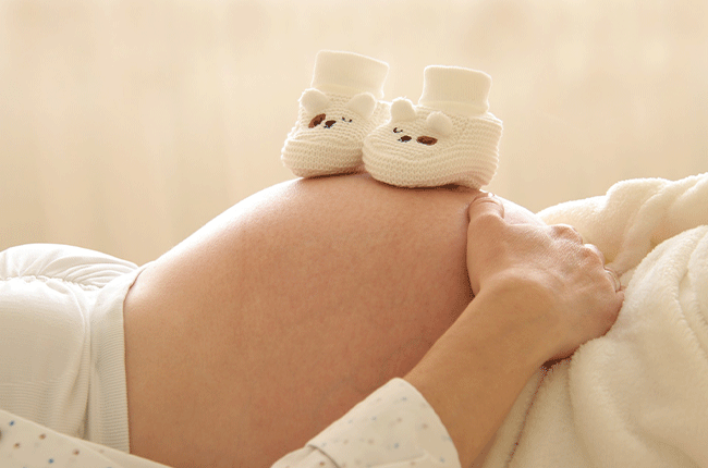 Bildausschnitt von dem Bauch einer liegenden Schwangeren. Darauf stehen kleine Stoffschuhe.