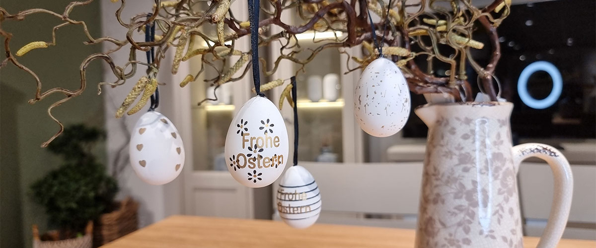 Auf einem Tisch steht eine Vase mit Zweigen. An ihnen hängen weiße Ostereier mit Motiven und Schriftzügen wie "Frohe Ostern".