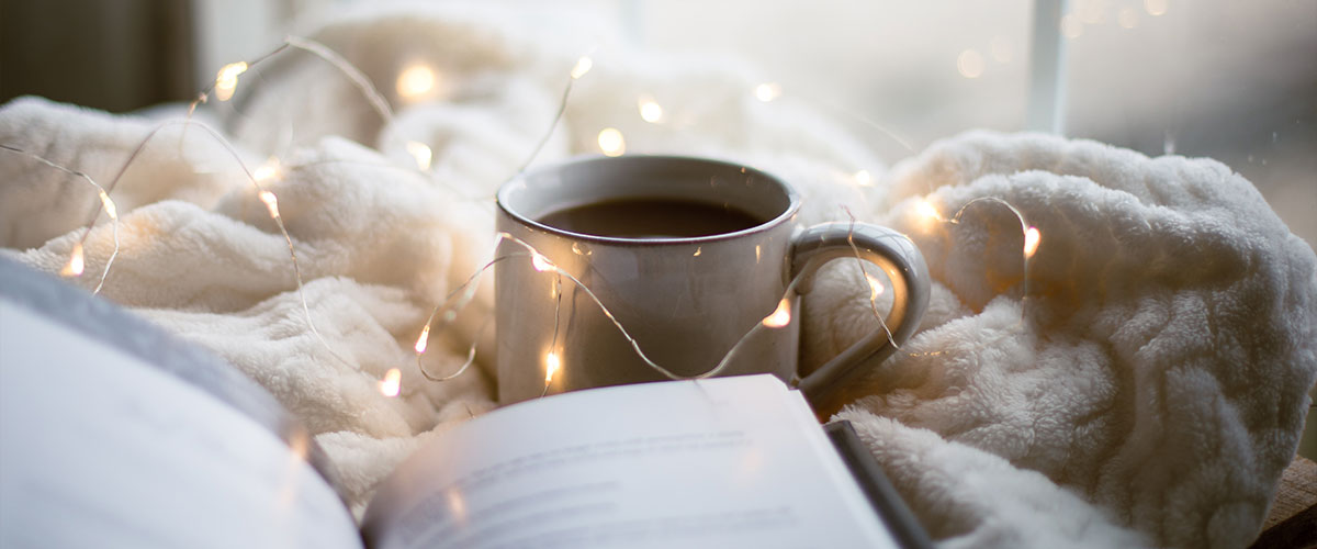 Eine Kaffeetasse auf einer Decke mit einer Lichterkette und einem Buch erzeugt Gemütlichkeit im Winter.