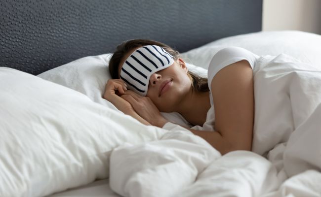 Eine Frau mit Schlafbrille liegt entspannt auf ihrem Kopfkissen im Bett.