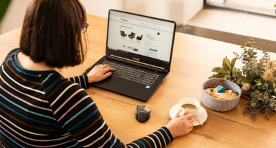 Eine Frau sitzt mit einem Laptop und einer Tasse Kaffee am Tisch ud guckt sich drei Stühle im Möbel Heinrich Online-Shop an.