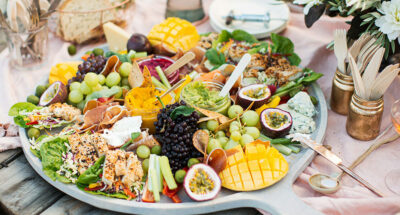 Ein Partybuffet mit einer Platte mit Obst, Dipps Gemüse, im Hintergrund Brot, Besteck ein Glas Sekt.