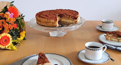 Ein gedeckter Kaffeetisch mit einer Eiskaffee-Torte, zwei Tellern mit Kuchenstücken, zwei Tassen mit Kaffee und einem sommerlichen Blumenstrauß.