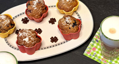 Ein Teller mit fünf Muffins und Schokoladen-Glücksklee, ein Glas Latte Macchiato auf einer Serviette mit Blumen.