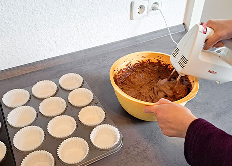 Zubereitung von Muffins für die Osterkaffeetafel: Muffinförmchen, eine Rührschüssel mit Teig, der mit einem Handrührgerät gerührt wird.