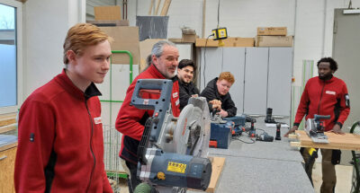 Mehrere Männer, die eine Ausbildung in der Möbelauslieferung machen, stehen in einer Werkstatt an der Werkbank, auf der mehrere Werkzeuge liegen.