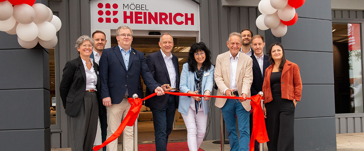 Eine Gruppe Frauen und Männer stehen vor dem Möbel Heinrich Eingang an einem roten Band. Drei Personen, schneiden das Band mit goldenen Scheren durch.