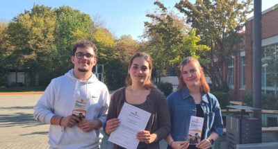 Zwei Schülerinnen und ein Schüler präsentieren den Ferienpass auf dem Schulhof.